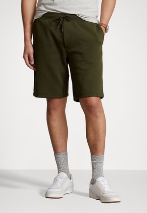 Shorts de survêtement vert olive avec une ceinture élastique, un cordon de serrage, des poches latérales et une coupe décontractée. Portés avec des chaussures blanches et des chaussettes grises.