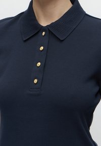Polo bleu marine avec col côtelé et quatre boutons dorés sur le devant, porté par une personne visible du cou jusqu'au milieu du torse.