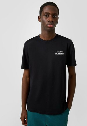 Jeune homme portant un t-shirt noir avec le texte blanc "INTO BRAZIL" sur la poitrine, un pantalon bleu sarcelle et des boucles d’oreilles en argent en forme de créoles.