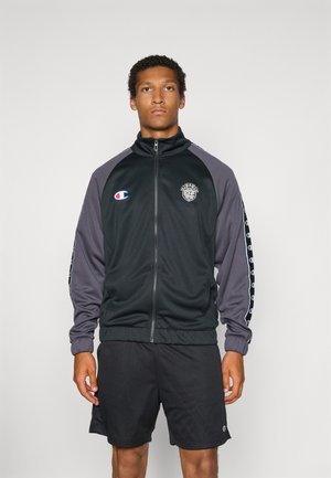 Ung mand iført en sort og grå sportsjakke med lynlås og logoer samt sorte meshshorts, stående foran en ensfarvet baggrund.