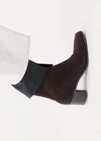 Καφέ μποτάκι αστραγάλου από σουέτ με τετράγωνη μύτη, υφή και κοντό τακούνι τύπου block, φορεμένο με μαύρη και λευκή ένδυση.