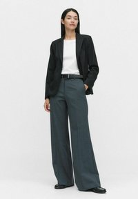 Schwarzer maßgeschneiderter Blazer über einem weißen Hemd, kombiniert mit grauen, weiten Hosen und einem schwarzen Gürtel. Strukturierter Stoff und lässige Passform.