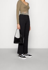 Kvinna i olivgrön rynkad topp med spetsdetaljer, svarta högmidjade utsvängda jeans, svartvita sneakers, håller i en svart texturerad handväska.