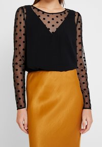 Černé polka dot průhledné dlouhé rukávy s pevným černým tělem, doplněné saténovou sukní ve zlatě žluté barvě. Hladká textura a přiléhavý střih.