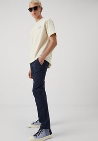 Beige short-sleeve t-shirt with embroidered logo, paired with dark navy slim-fit pants and two-tone high-top sneakers. Black sunglasses worn.
