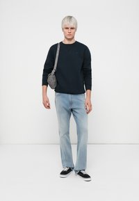 Felpa crewneck blu navy, jeans dritti azzurri chiari, sneaker nere e una borsa a tracolla grigia con una tracolla caratterizzata da accessori in ottone argentato.