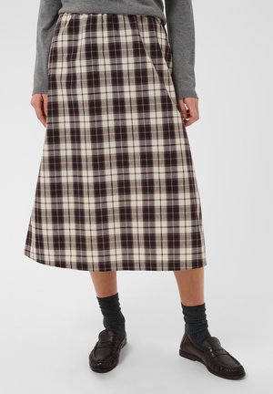 Femme portant une jupe midi à carreaux marron et crème, un pull gris, des chaussettes gris foncé et des chaussures marron foncé à enfiler, debout sur un fond blanc.