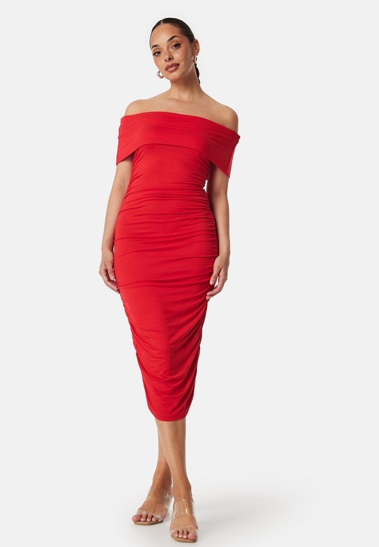 Robe rouge à épaules dénudées avec une silhouette ajustée, texture froncée et ourlet au genou. Des bretelles transparentes et des sandales couleur chair complètent le look.