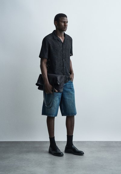 Άνδρας που φοράει μαύρο δαντελένιο πουκάμισο με κοντά μανίκια, τζιν σορτς, μαύρες κάλτσες και παπούτσια, κρατώντας μια μεγάλη μαύρη δερμάτινη τσάντα.