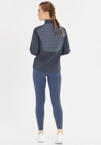 Kvinna med flätat hår som bär en mörkgrå jacka och blå leggings, står vänd bort från kameran mot en helt vit bakgrund.