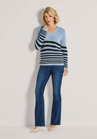 Blonde Frau, die einen hellblauen und schwarzen gestreiften Strickpullover, dunkelblaue Schlagjeans und beige High Heels trägt, steht vor einer neutralen Wand.