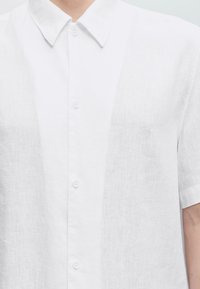 Gros plan d'une personne portant une chemise blanche en lin à manches courtes par-dessus une chemise blanche à col boutonné.