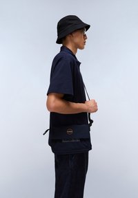 Marinblå kortärmad polo tröja i kombination med en svart bucket hat och en marinblå crossbody-väska med en cirkulär logotyp och rem.