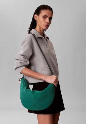 Žena v sivej pulóvri a čiernej sukni drží zelenú polmesiacovú kabelku s ramenným popruhom na sivom pozadí.