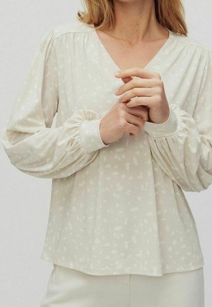 Femme portant un chemisier beige clair à col en V avec de subtiles taches blanches et des manches longues bouffantes avec poignets ajustés.