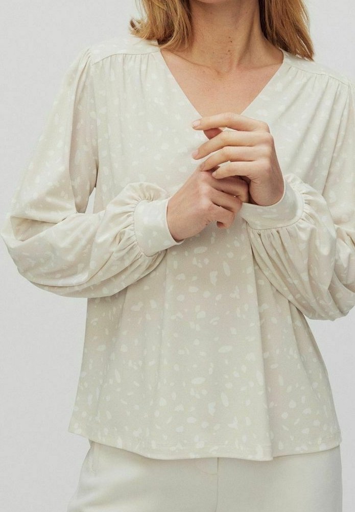 Femme portant un chemisier beige clair à col en V avec de subtiles taches blanches et des manches longues bouffantes avec poignets ajustés.