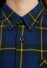 Camicia di flanella con un motivo a quadri blu, verde e giallo, colletto a punta con chiusure a bottone e taschino sul lato sinistro.
