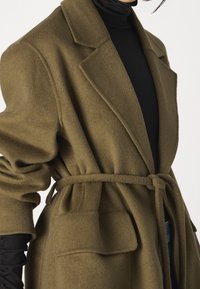 Oliivinvihreä, pehmeä kangastakki, jossa on kaulus, pitkät hihat ja solmittava vyötärövyö. Takissa on kaksi etutaskua ja rento istuvuus.