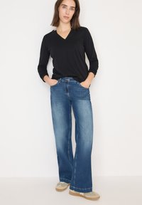 Schwarzes langärmliges V-Ausschnitt-Shirt kombiniert mit hellblauen weit geschnittenen Jeans, ausgestattet mit Fronttaschen und braunem Schuhwerk mit grauen Akzenten.