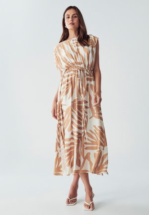 Maxi dress in lightweight fabric with a beige and white leaf pattern, elasticized waist, and one-shoulder design. Worn with white sandals.