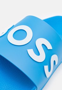 Modré gumové sandály s kluzkým designem, které mají velké embosované bílé logo. Texturovaný interiér pro lepší grip, minimalistický otevřený design na špičce.