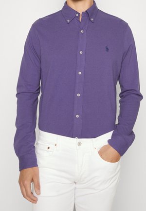 Mann trägt ein violettes Hemd mit langen Ärmeln und Knöpfen, mit einem kleinen gestickten Logo auf der Brust und weiße Hose, eine Hand in der Tasche.