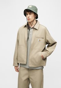 Mladý muž v béžovej bunde a nohaviciach, pruhovanej košeli a zelenej klobúku s remienkom pod bradou, stojaci na neutrálnom pozadí.