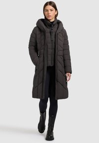 Mujer joven vestida con un abrigo largo negro acolchado sobre una chaqueta negra con cremallera, leggings negros y botas negras hasta el tobillo, de pie con las manos en los bolsillos.