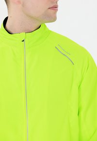 Vihreä neon-väri taitettava takki korkealla kauluksella, jossa on heijastavat yksityiskohdat ja sileä pinta; logo "ENDURANCE" olkapäällä.