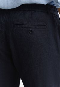 Gros plan sur un pantalon en lin bleu marine foncé avec une poche arrière boutonnée, porté avec une chemise bleu clair rentrée.
