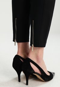 Zwarte tapered broek met enkelritsen en een gladde afwerking, gecombineerd met zwarte suède hakken met een spitse neus en een open zijkant.