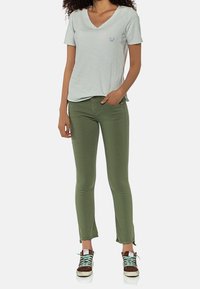 Camiseta verde claro de cuello en V con un pequeño logo azul, combinada con pantalones ajustados verde oliva y zapatillas marrones con cordones turquesa.