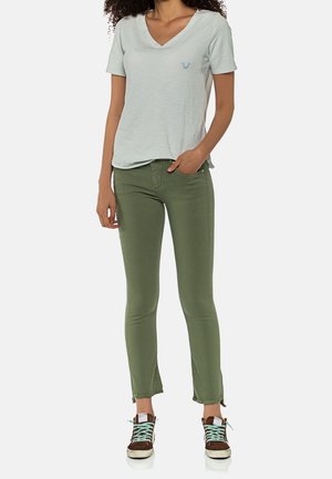 Camiseta verde claro de cuello en V con un pequeño logo azul, combinada con pantalones ajustados verde oliva y zapatillas marrones con cordones turquesa.