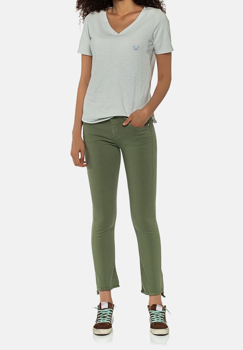 Camiseta verde claro de cuello en V con un pequeño logo azul, combinada con pantalones ajustados verde oliva y zapatillas marrones con cordones turquesa.