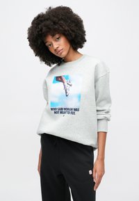 Světlé šedé tričko s grafickým potiskem nohy v teniskách na pozadí modré oblohy. Černé kalhoty s malým logem na straně.