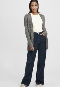 Grauer gestrickter Cardigan mit Knopfleiste, über einem hellen cremfarbenen Oberteil und einer dunkelblauen Corduroy-Hose in weitem Schnitt getragen. Strukturierter Stoff, bequeme Passform.