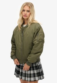 Oliwkowa kurtka bomberka z zamkiem z przodu, ściągaczami przy rękawach oraz kieszenią na lewym rękawie, zestawiona z czarno-białą spódnicą w kratę.