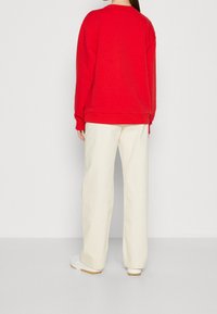 Roter Sweatshirt mit gerippten Bündchen und Saum, kombiniert mit cremefarbenen, weit geschnittenen Hosen und weißen Turnschuhen, von hinten gesehen.