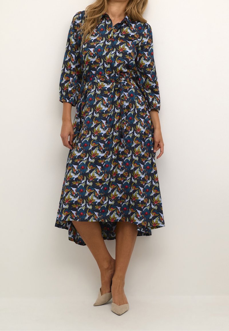 Femme portant une robe midi fleurie bleu marine avec des manches trois-quarts, un col, et des talons beiges à bouts pointus, debout devant un fond uni.