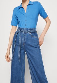 Blue short-sleeve polo shirt with a button placket paired with high-waisted, wide-leg denim trousers featuring a tied belt and front pockets.