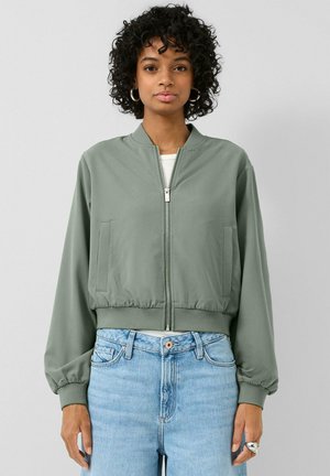 Femme aux cheveux courts et bouclés portant une veste bomber zippée vert clair, un jean bleu clair, des boucles d'oreilles anneaux et une bague argentée, debout devant un fond uni.