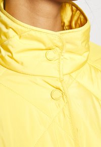 Gelbe gesteppte Jacke mit hohem Kragen, ausgestattet mit zwei runden Knöpfen und einer glatten, glänzenden Stoffoberfläche.