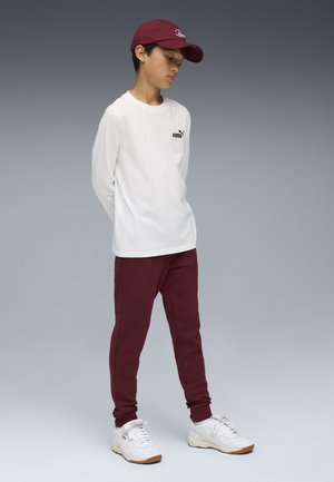 Burgunderfarvede sweatpants, lang hvid t-shirt med en lille sort logo, og hvide sportssko. En burgunderfarvet kasket fuldender outfittet.