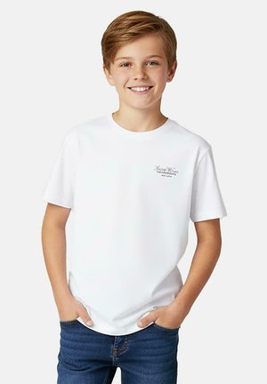 Lachende jongen met kort lichtbruin haar, gekleed in een wit T-shirt met korte mouwen en blauwe spijkerbroek, handen in de zakken, staand tegen een witte achtergrond.