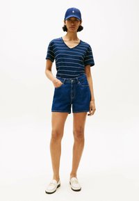 Camiseta de rayas azules con escote en V, combinada con pantalones cortos de denim de tiro alto. El modelo lleva una gorra azul y loafers blancos con acentos dorados.