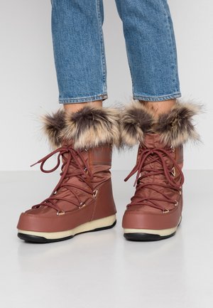 Botas de invierno marrones con borde de piel y cordones, usadas con vaqueros azules, mostradas desde la mitad de la pantorrilla hacia abajo sobre un fondo blanco.