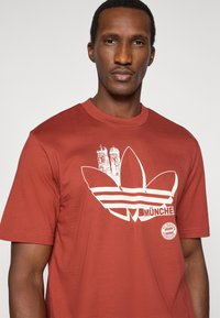 Μακρυμάνικη κόκκινη μπλούζα από βαμβάκι με μεγάλο λευκό λογότυπο Adidas trefoil με τρεις ρίγες, διακοσμημένη με γραφικά του Μονάχου και σήμα της FC Bayern.