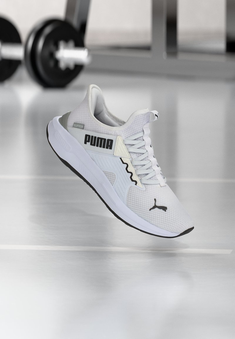 Λευκό αθλητικό παπούτσι Puma με μαύρο λογότυπο και ανώτερο μέρος από διχτυωτό υλικό, αιωρούμενο πάνω από μια αντανακλαστική γυμναστηριακή επιφάνεια με ένα μπάρα βαρών στο θολωμένο φόντο.