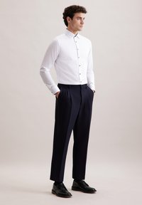 Camicia bianca a maniche lunghe con bottoni neri, abbinata a pantaloni blu con pieghe e scarpe nere, il tutto su uno sfondo neutro.