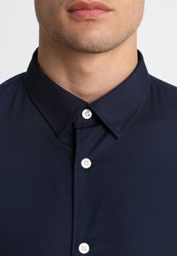 Gros plan sur la partie inférieure du visage et le cou d’un homme portant une chemise bleu marine à boutons blancs et col pointu.